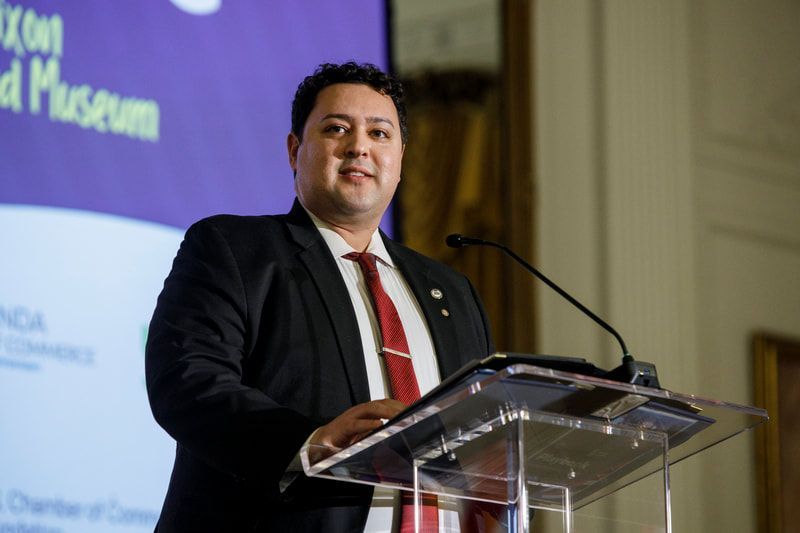 Man speaking at a podium, wearing a suit and tie, in front of a banner.