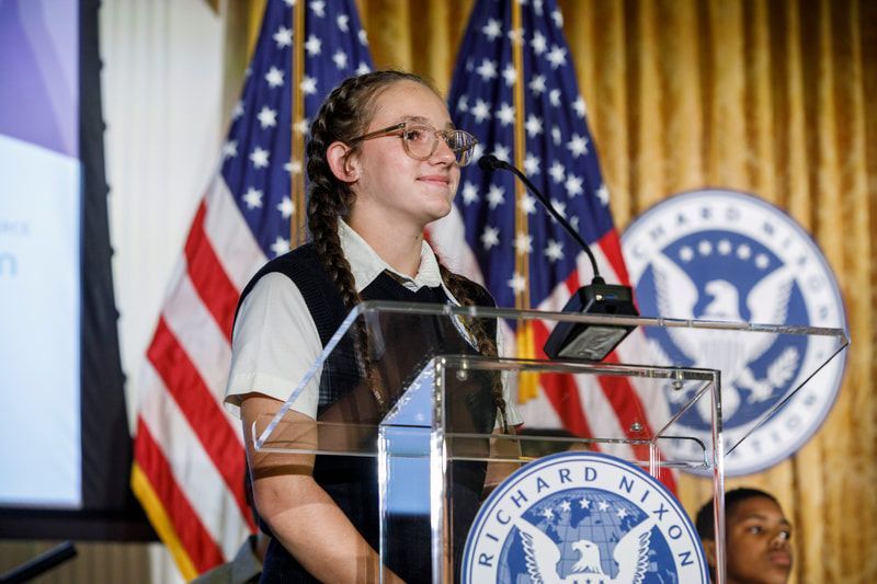 Girl speaking at a podium with American flags and a Nixon Presidential Library logo in the background.
