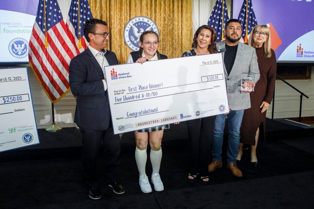 Group of people holding an oversized check indoors with flags and a screen.