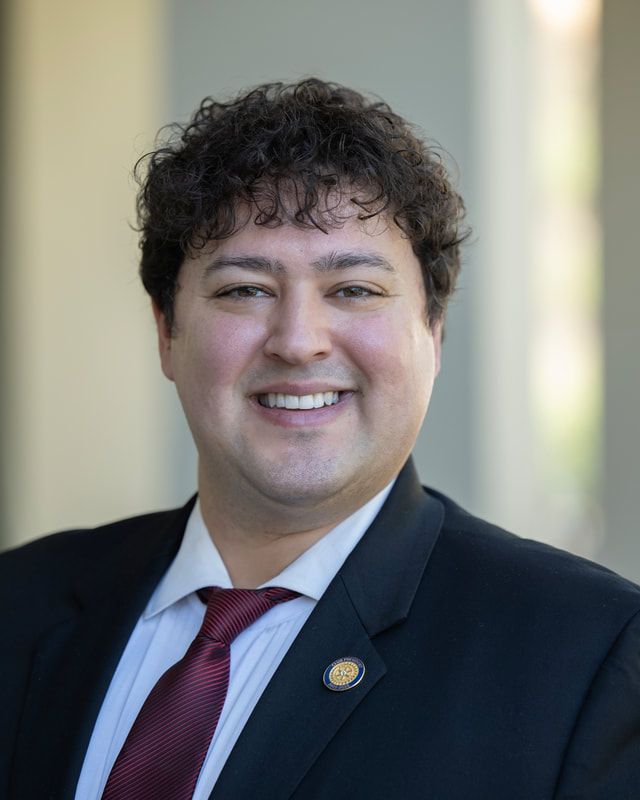 Man in a suit smiles, dark curly hair, red tie, and a lapel pin.