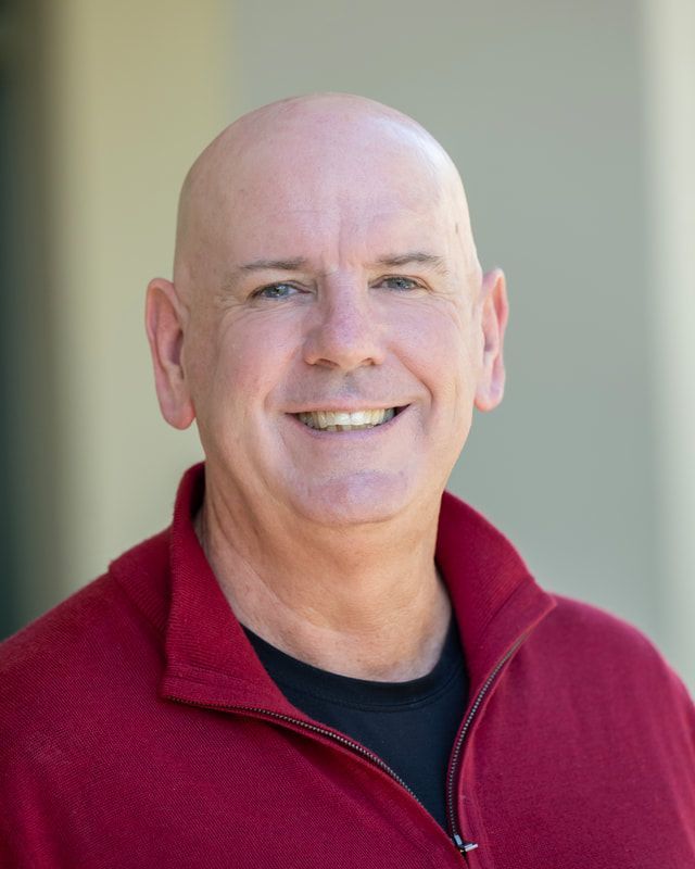 Bald man smiles, wearing a red zip-up jacket over a black shirt, outdoors.