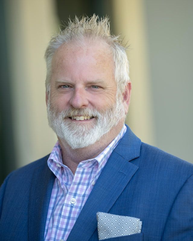 Man with graying hair and beard smiles, wearing a blue blazer and patterned shirt outside.