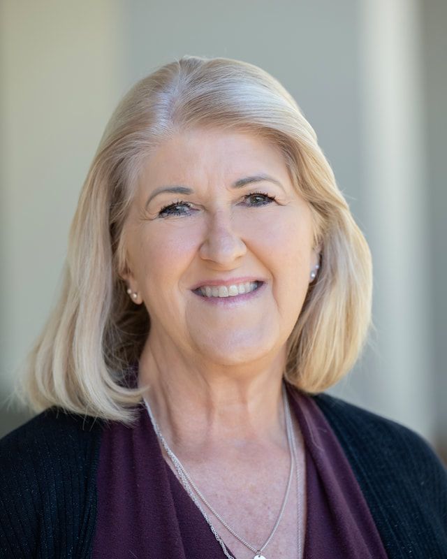 Woman with blonde hair smiles, wearing a black cardigan and maroon shirt, outdoors.