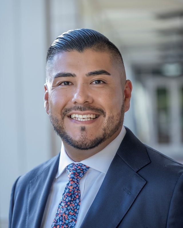 Man in a suit smiles at the camera, with a close shaved haircut and patterned tie.