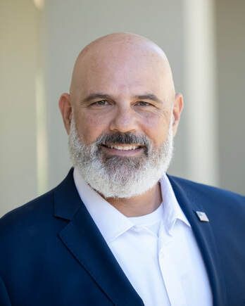 Bald man with gray beard, smiling, wearing a dark blue suit and white shirt, outdoors.