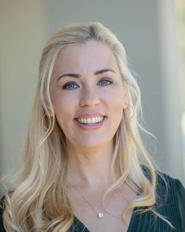 Woman with blonde hair smiles at the camera, wearing gold hoop earrings and a necklace; outdoors.