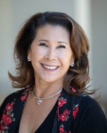 Woman with brown hair smiles, wearing floral blazer and silver necklace, outdoors.
