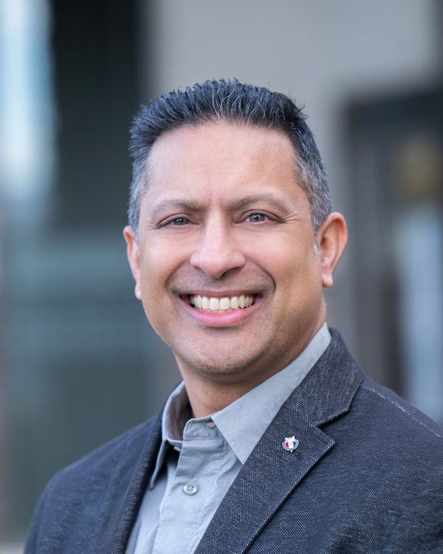Man with dark hair smiling, wearing a gray shirt and dark blazer, small pin on lapel; blurred background.