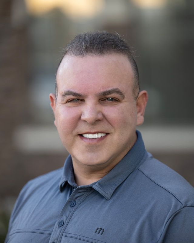 Man in a blue shirt smiles at the camera, outdoors in front of a building.