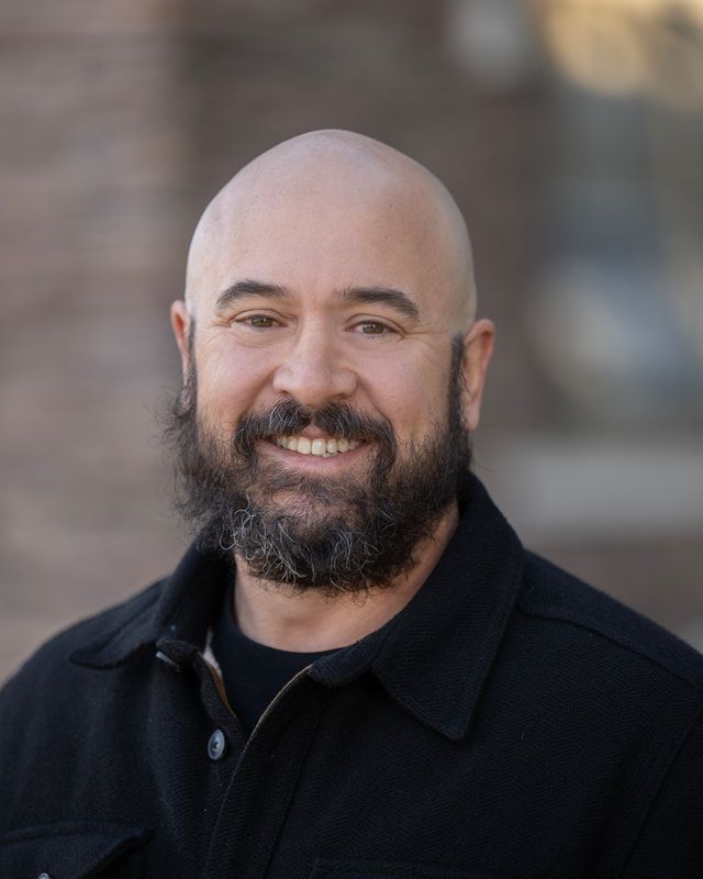 Bald man with a full beard smiles, wearing a black shirt, set against a blurred brick background.