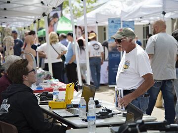 Man in white shirt talking to person at a table with equipment, outdoor event with tents and people.