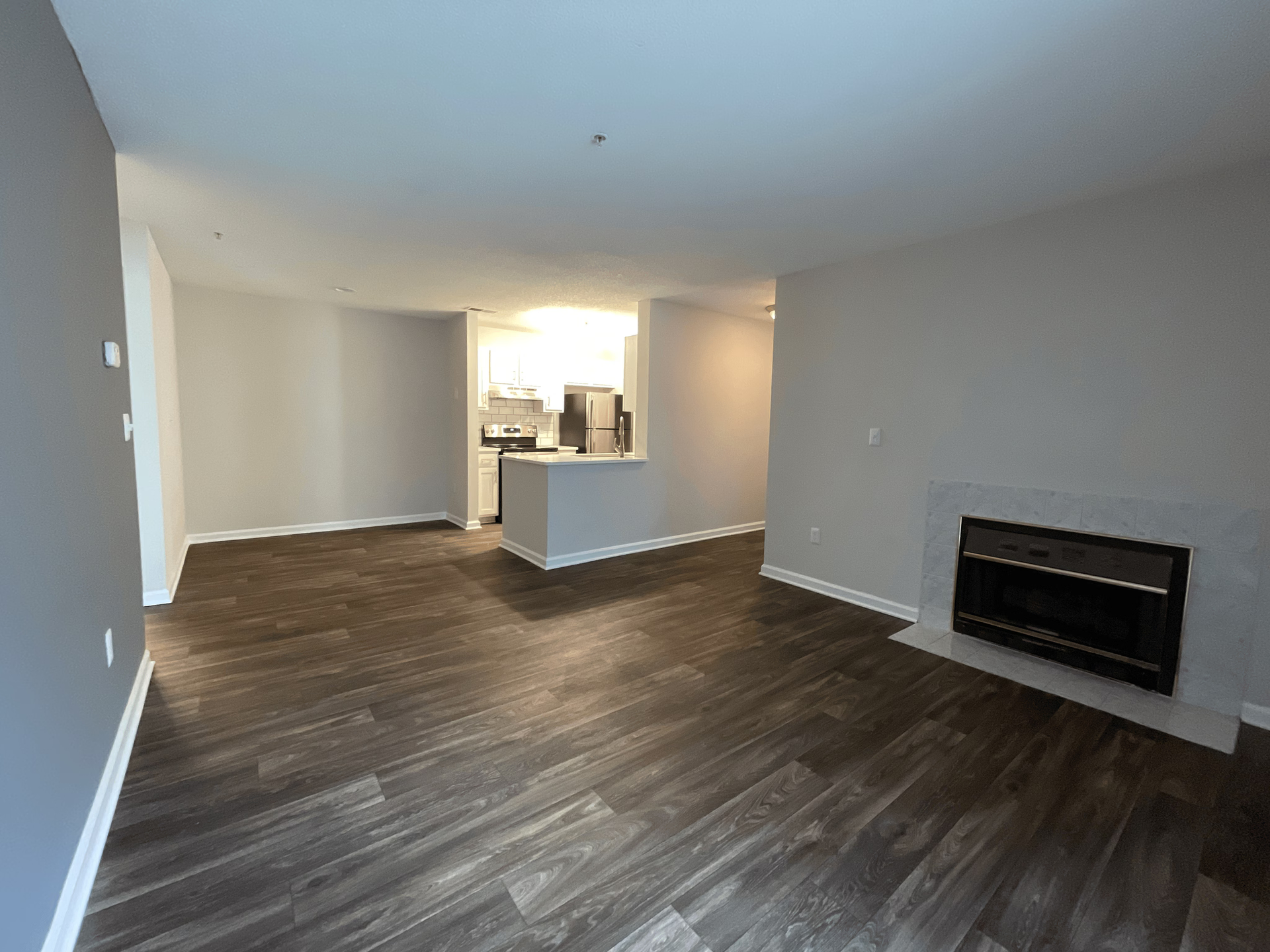 Empty apartment living room with fireplace, dark wood flooring, and gray walls. Kitchen visible in the background.