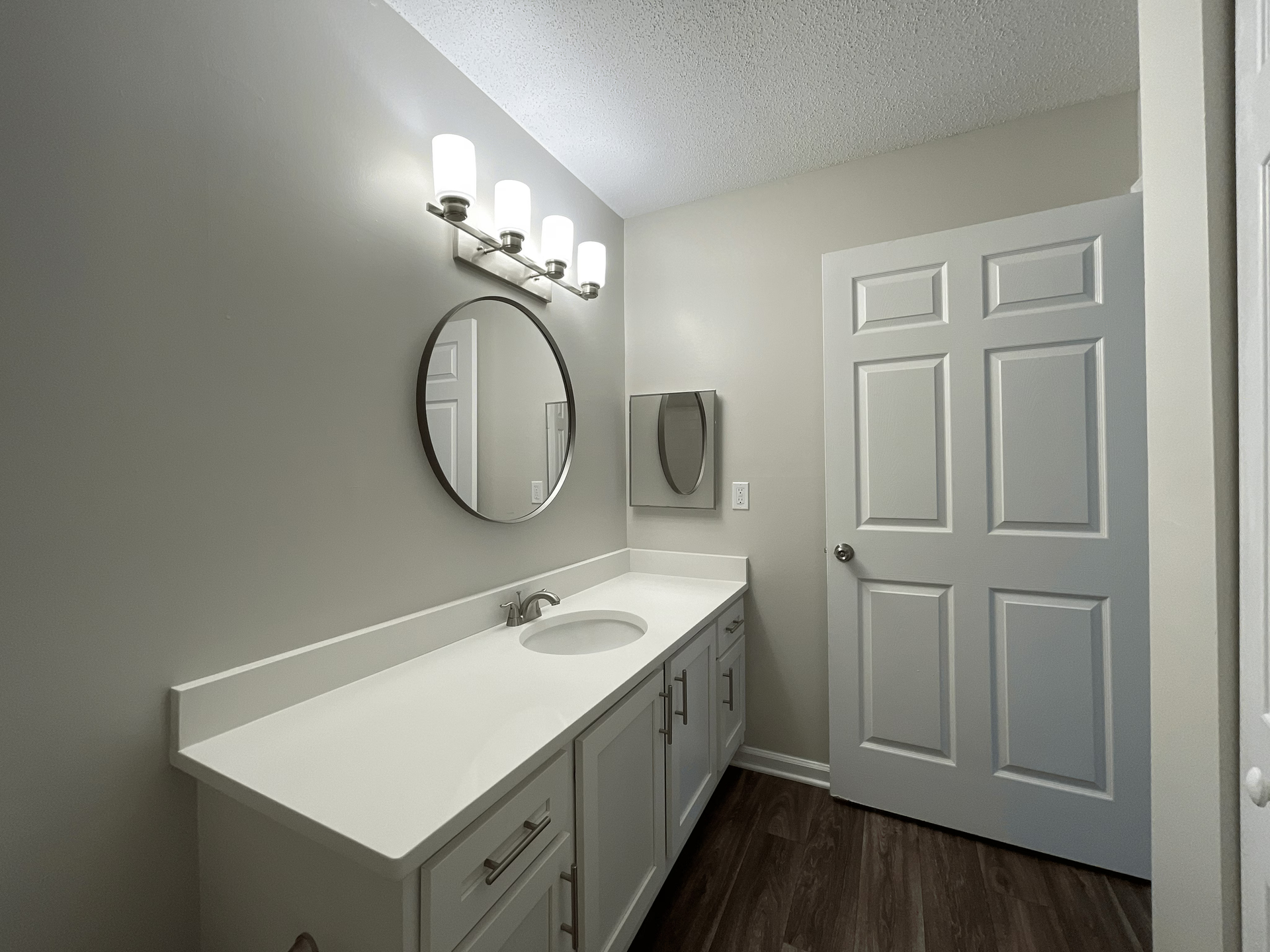 Bathroom with white countertop, round mirror, white cabinets, and a closed white door.