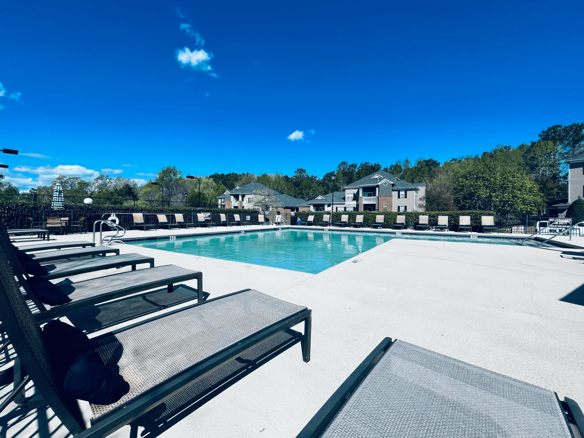 Swimming pool with lounge chairs on a sunny day. Apartment buildings in background. Blue sky.