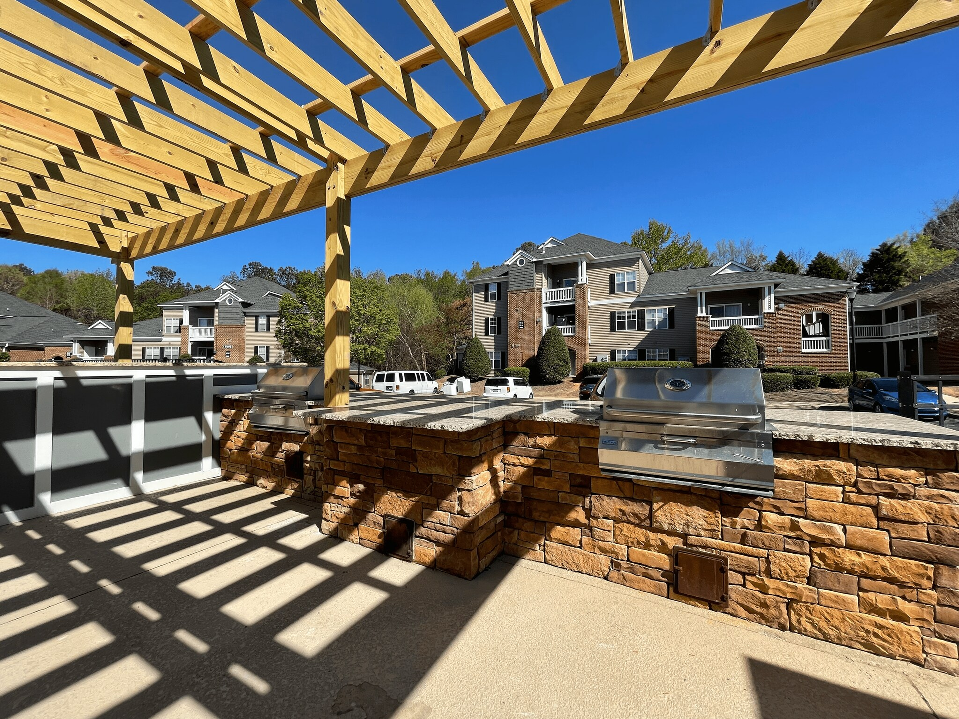 Outdoor grilling area with pergola, stone walls, and apartment buildings in background.