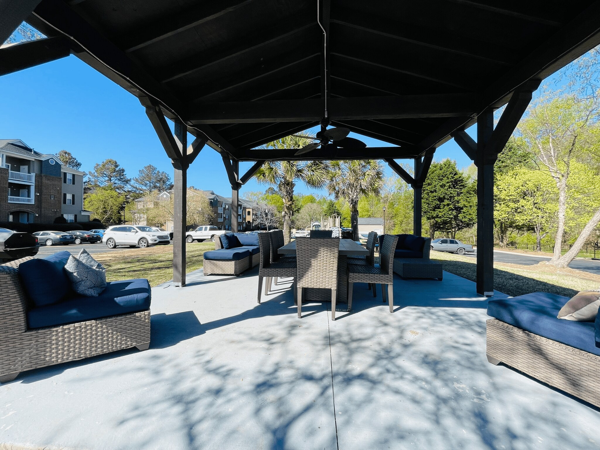 Outdoor gazebo with blue seating, table, and trees. Concrete floor and blue sky.