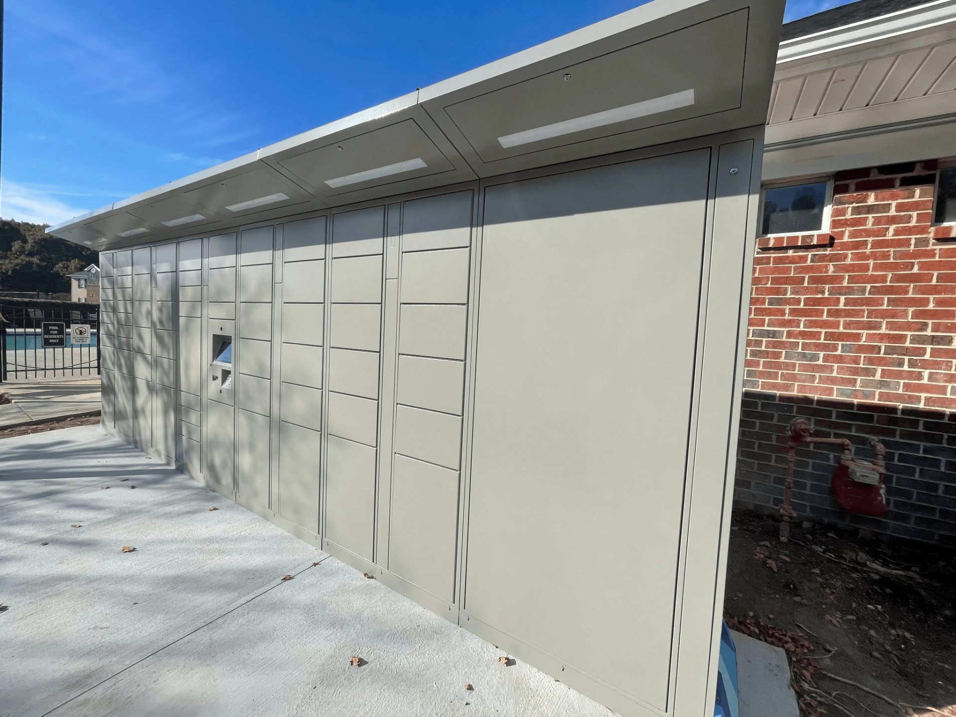 Gray parcel locker with slanted roof, near a brick building and a pool area.