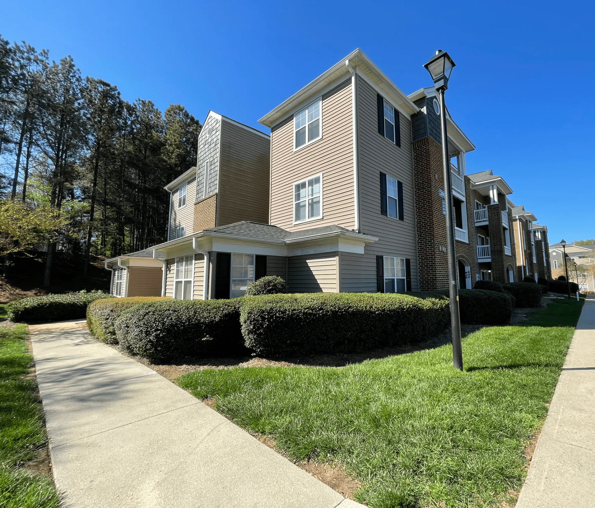 Multi-story beige apartment building with bushes, sidewalk, and street lamp.