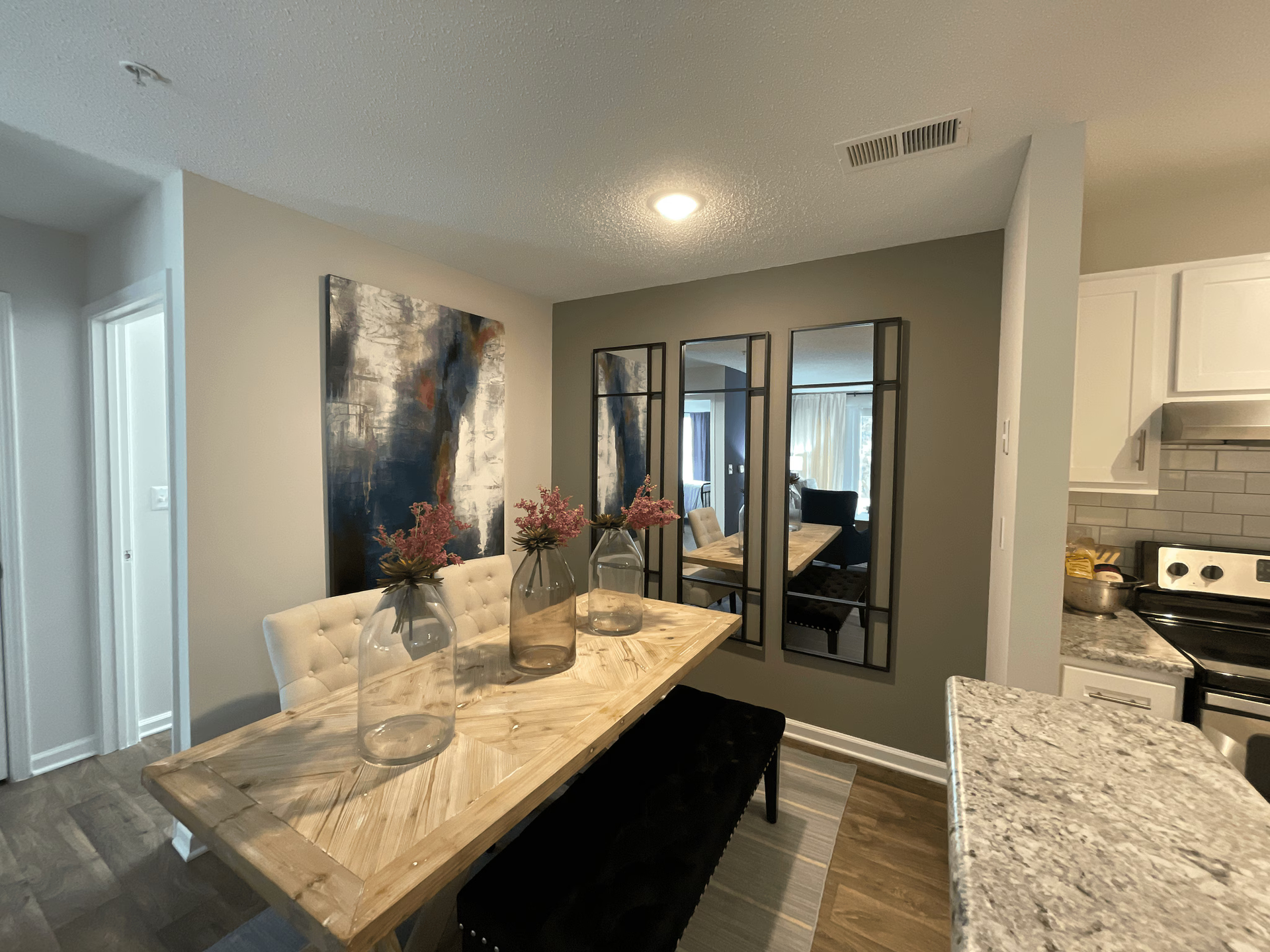 Dining room with table, bench, three mirrors, and artwork; a kitchen is partially visible.