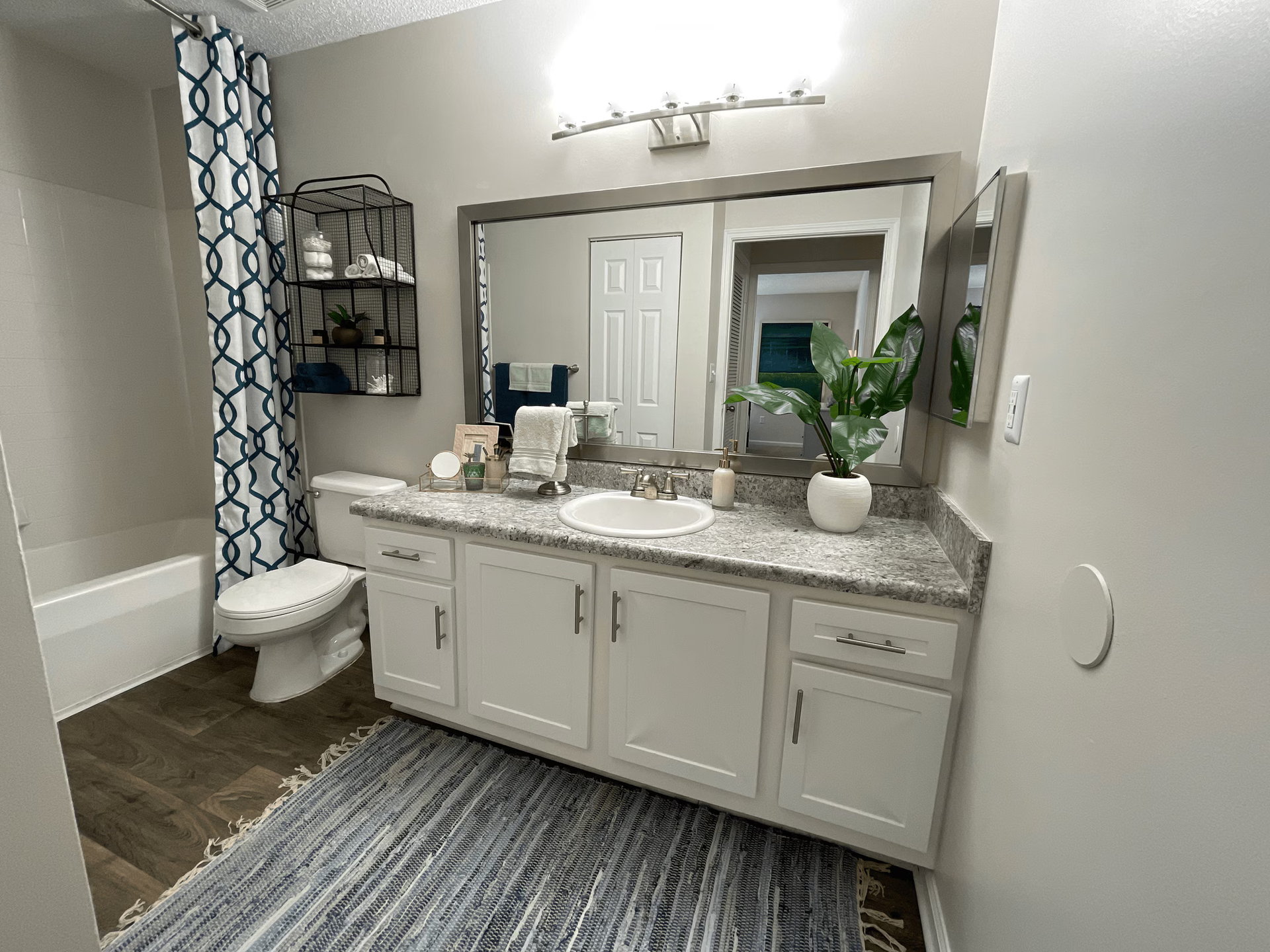 Bathroom with white cabinets, gray countertop, large mirror, patterned rug, and shower curtain.