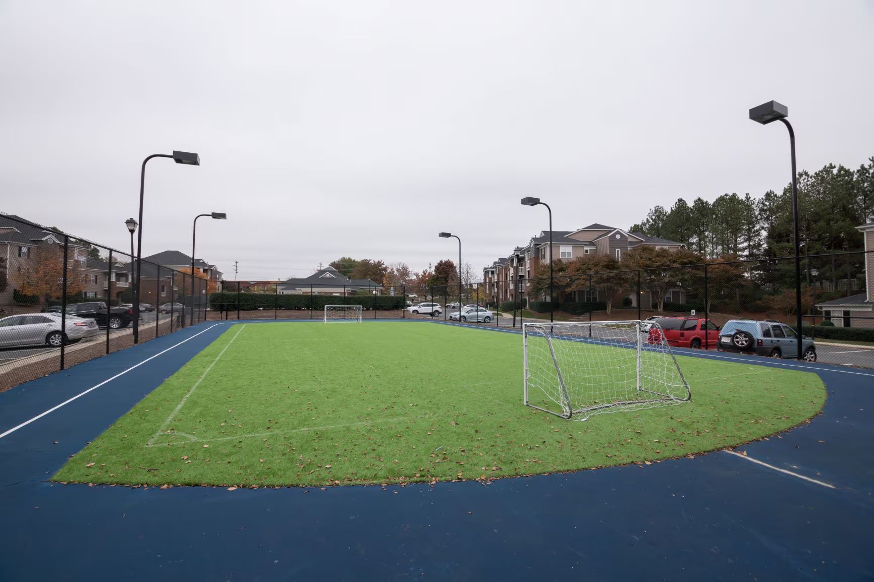 Soccer field with artificial turf, blue surrounding area, goal, and streetlights. Cars and buildings in the background under an overcast sky.