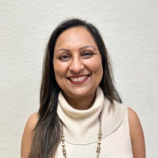 Woman with long dark hair smiles, wearing a cream-colored turtleneck and necklace against a white background.