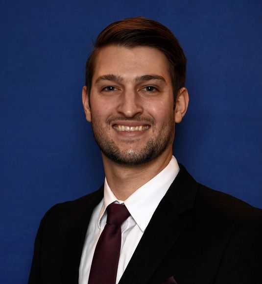 Man in a suit smiles at the camera against a blue background. He wears a tie.