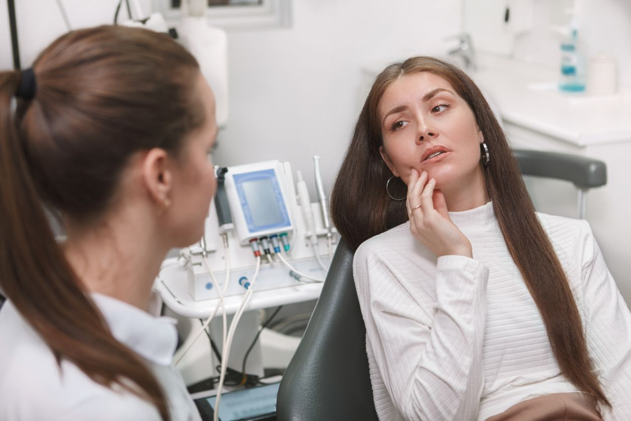 Woman in dental chair, holding jaw, talking with dentist, in examination room.