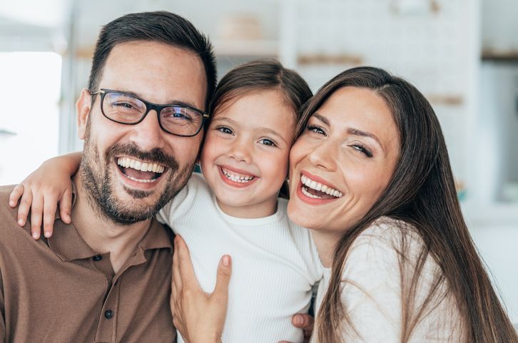 Family of three, smiling and hugging, in a bright home setting.