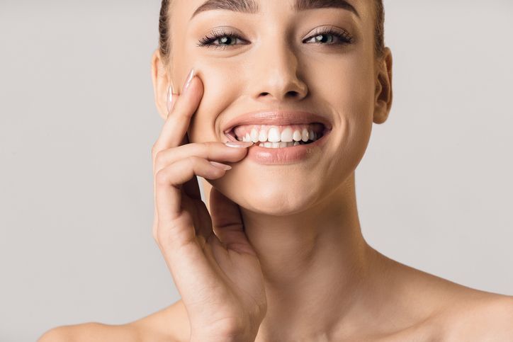 Woman smiling, touching cheek, showing teeth. Plain background.