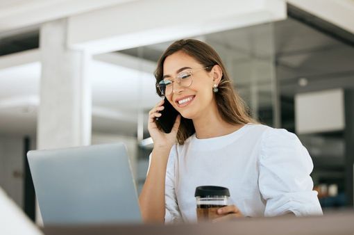 A woman is talking on a cell phone while using a laptop computer.