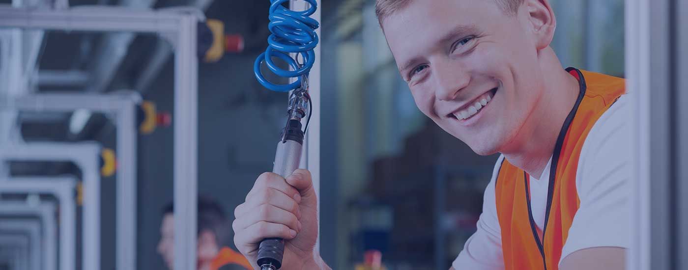 A man in an orange vest is smiling while holding a tool in a factory.