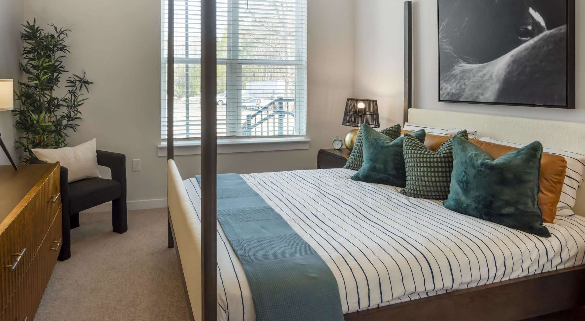 Cozy bedroom with a bed, window with blinds, dresser, plant, and bedside lamp.