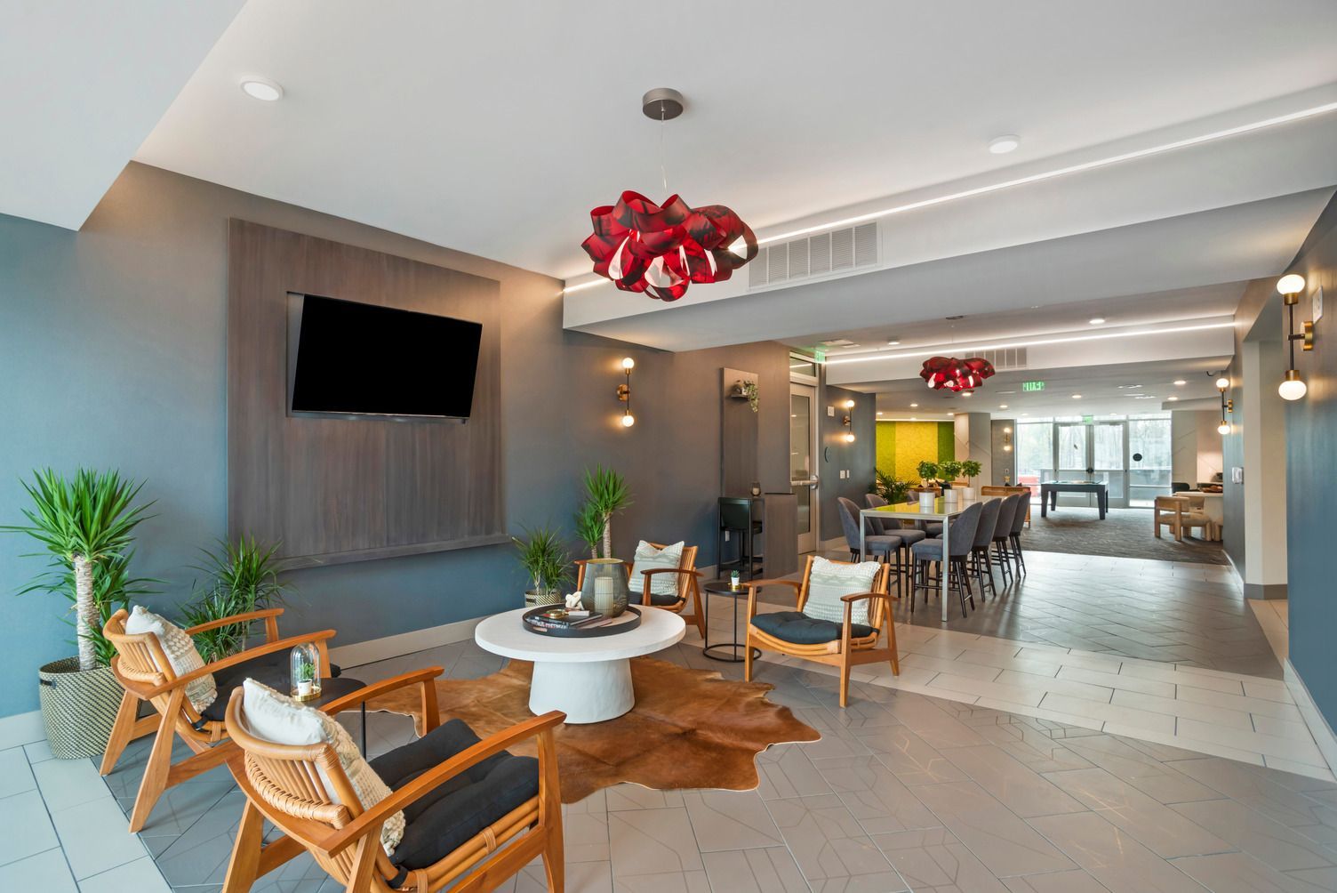 Spacious modern community lounge with seating, a wall-mounted TV, plants, and a dining area.