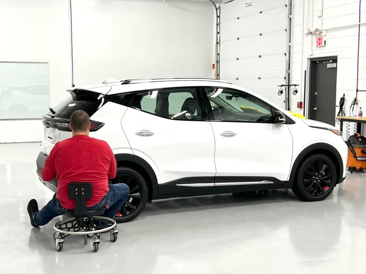 A man is sitting on a stool in front of a white car in a garage.