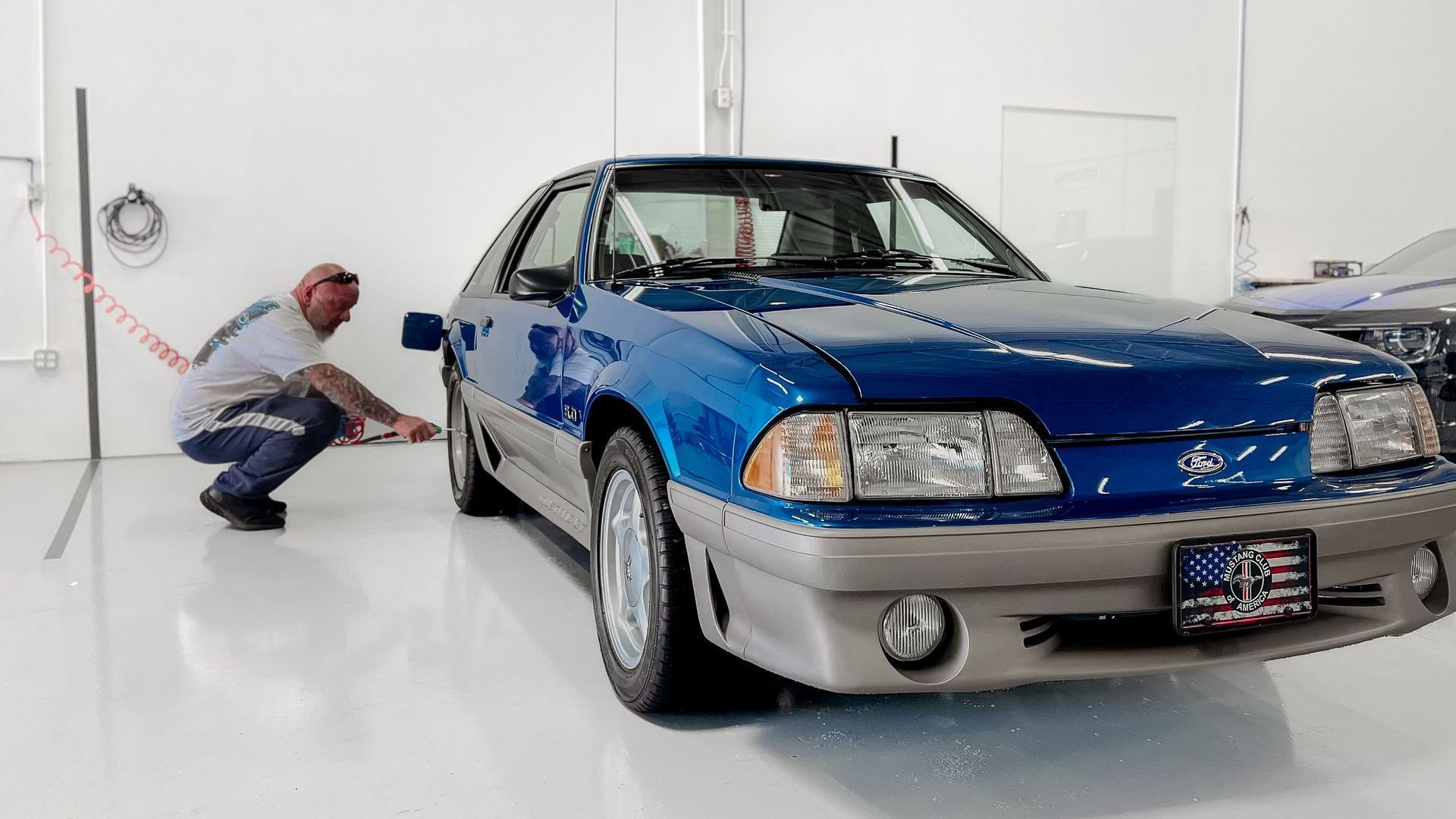 A man is squatting next to a blue mustang in a garage.