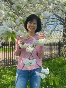 A woman in a pink sweater is standing in front of a tree with white flowers.