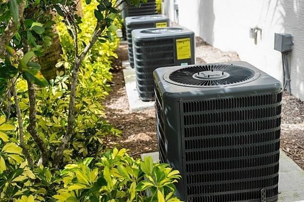 Row of black air conditioning units outside, next to green foliage and a white wall.