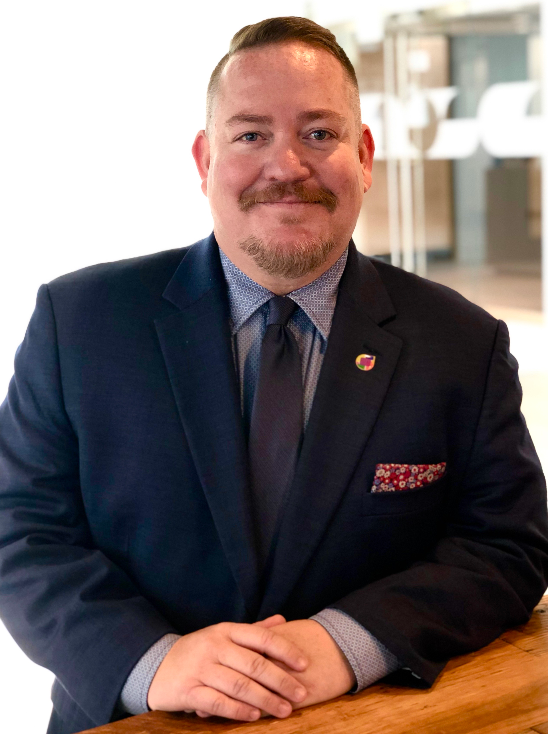 A man in a suit and tie is sitting at a table with his hands folded.