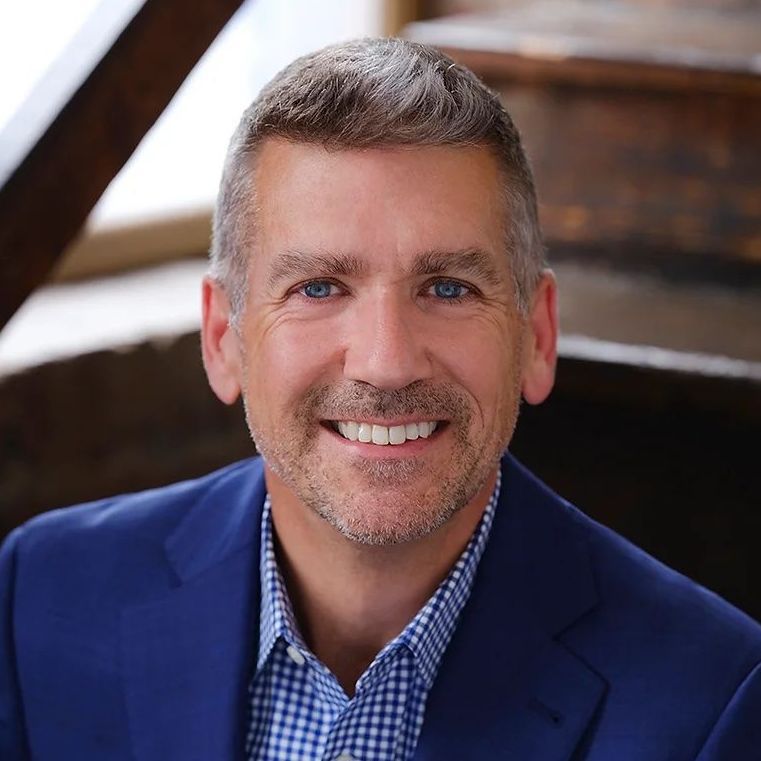 A man in a blue suit and plaid shirt is smiling for the camera.
