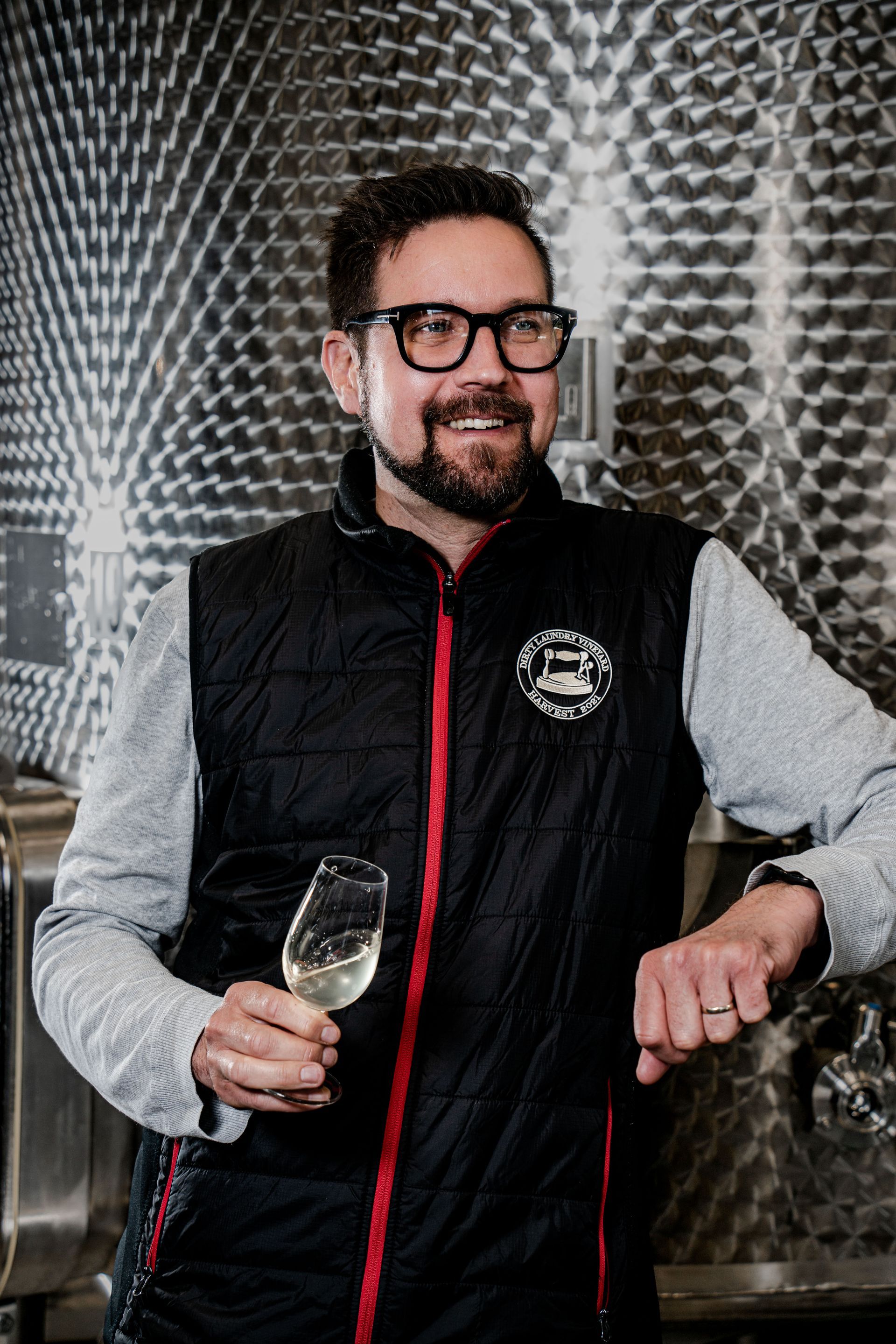 A man with a beard and glasses is holding a glass of wine.