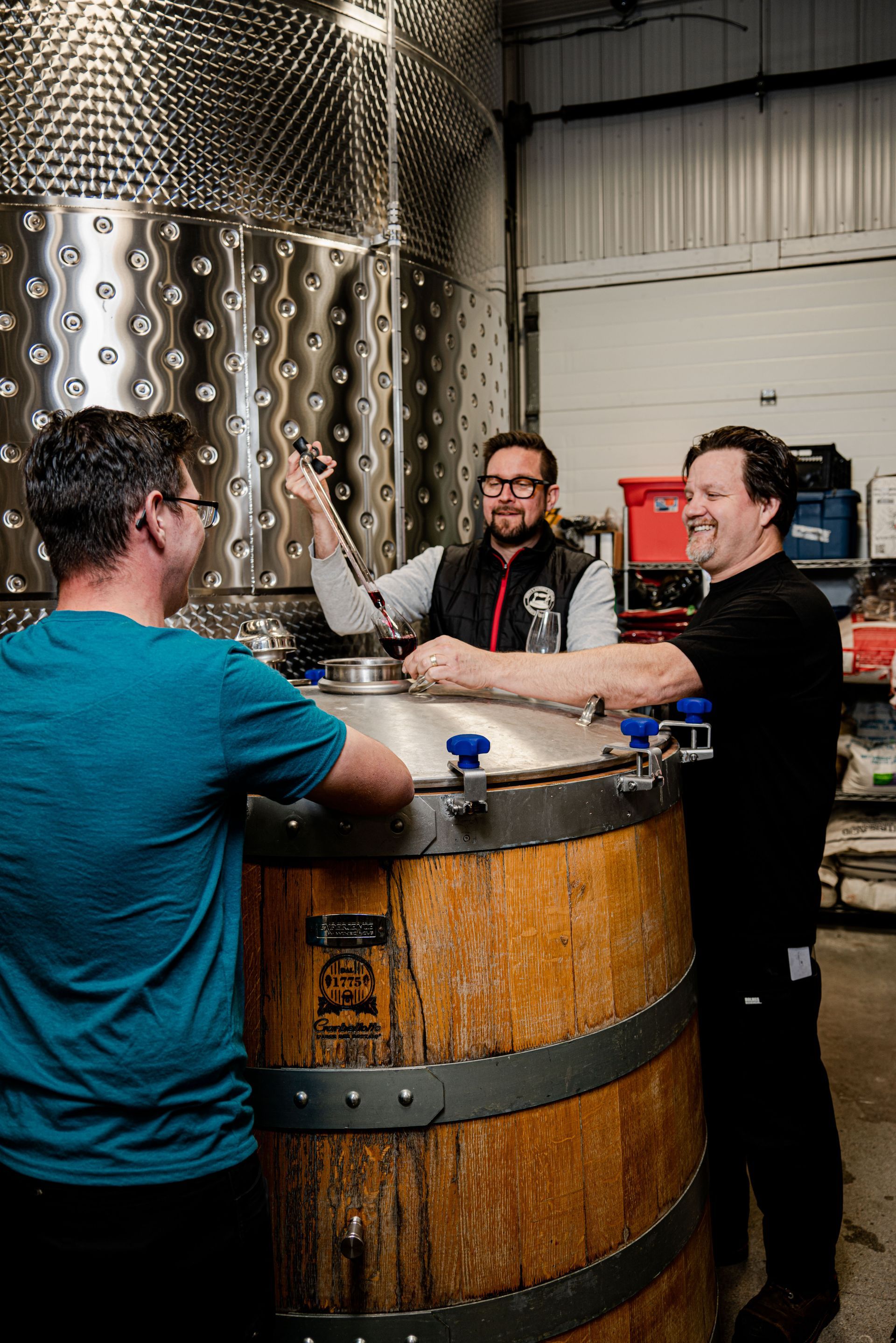 Three men are standing around a wooden barrel in a warehouse.