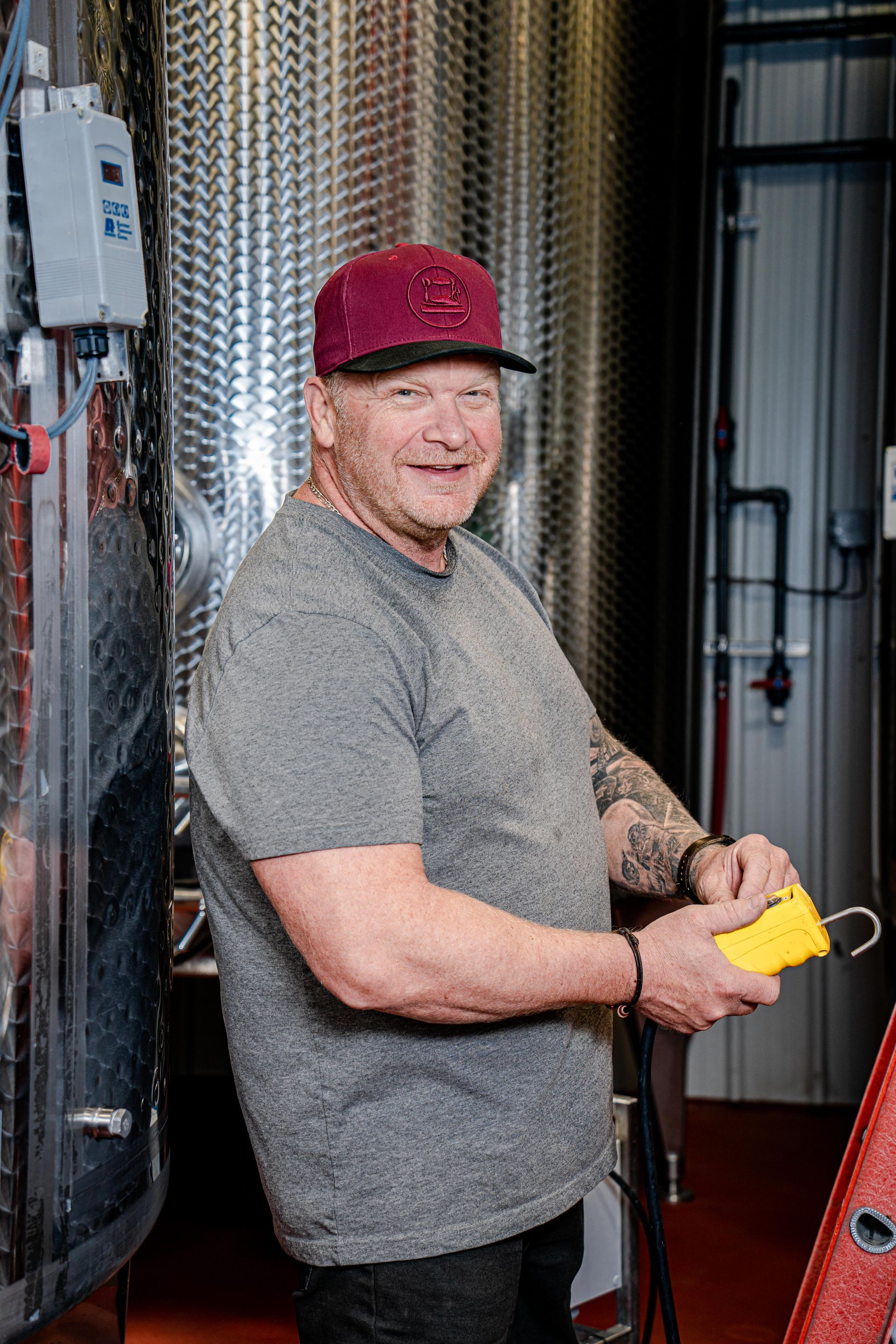A man wearing a hat and a gray shirt is standing in front of a stainless steel tank.