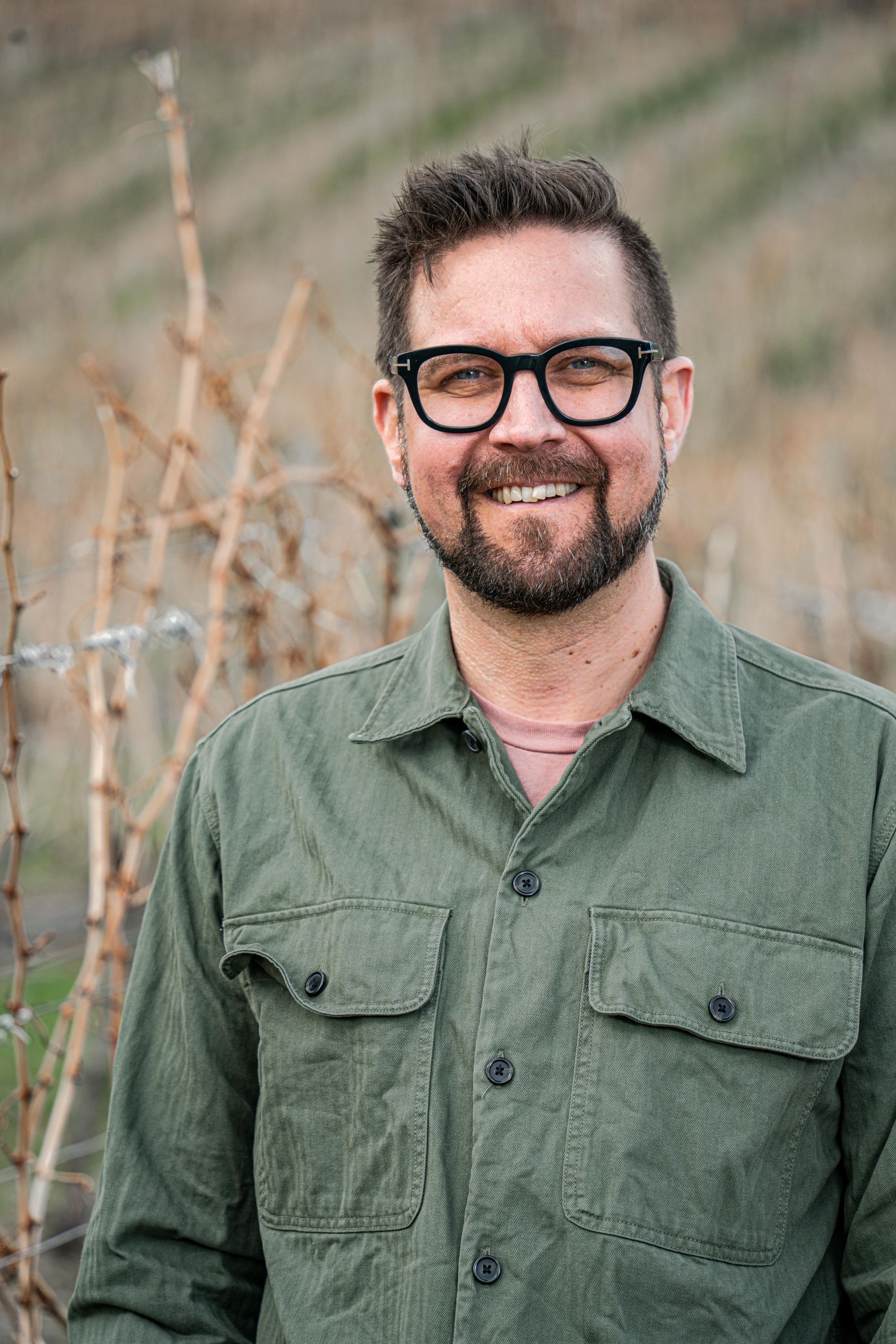 A man with a beard wearing glasses and a green shirt is standing in a vineyard.