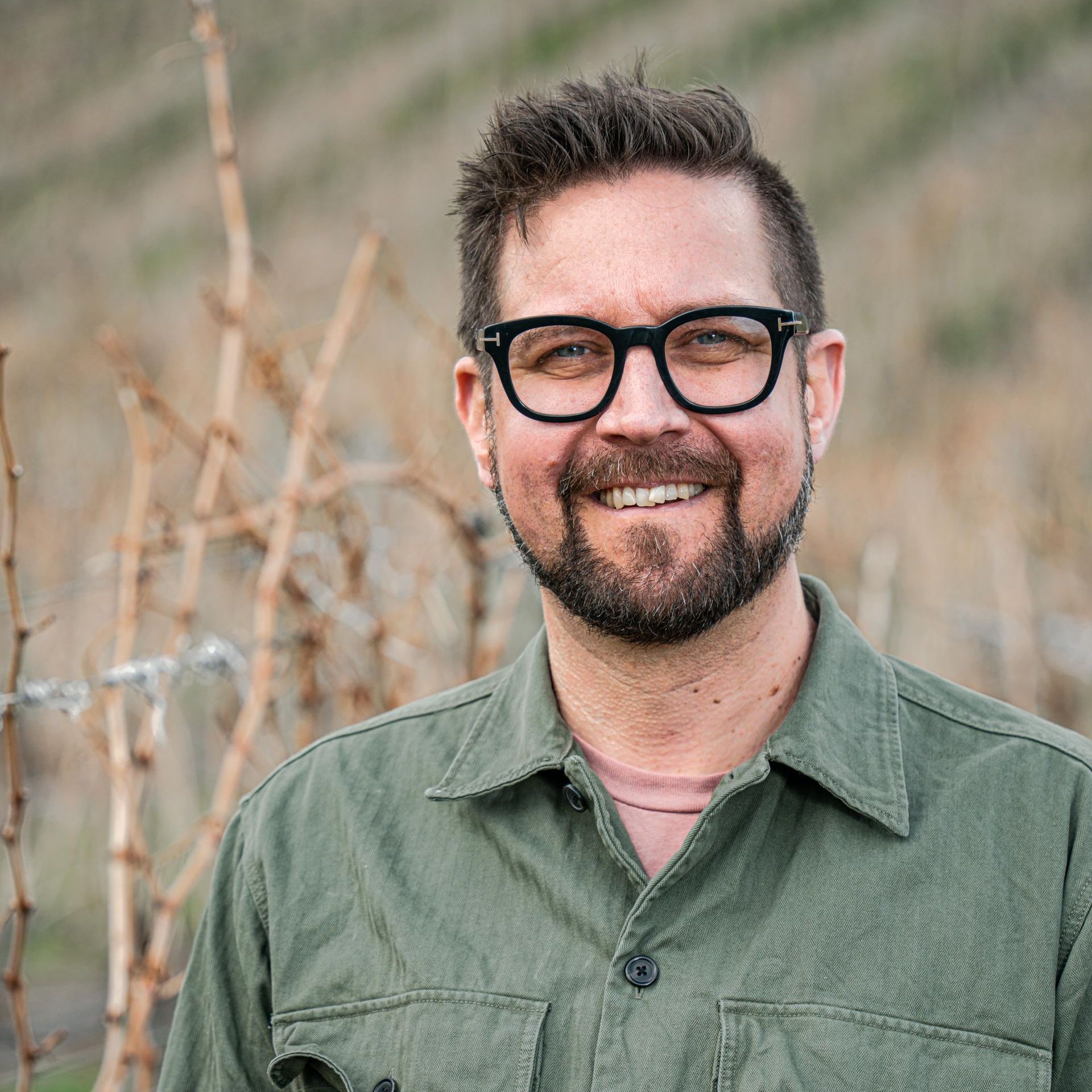 A man with a beard and glasses is smiling in a vineyard.