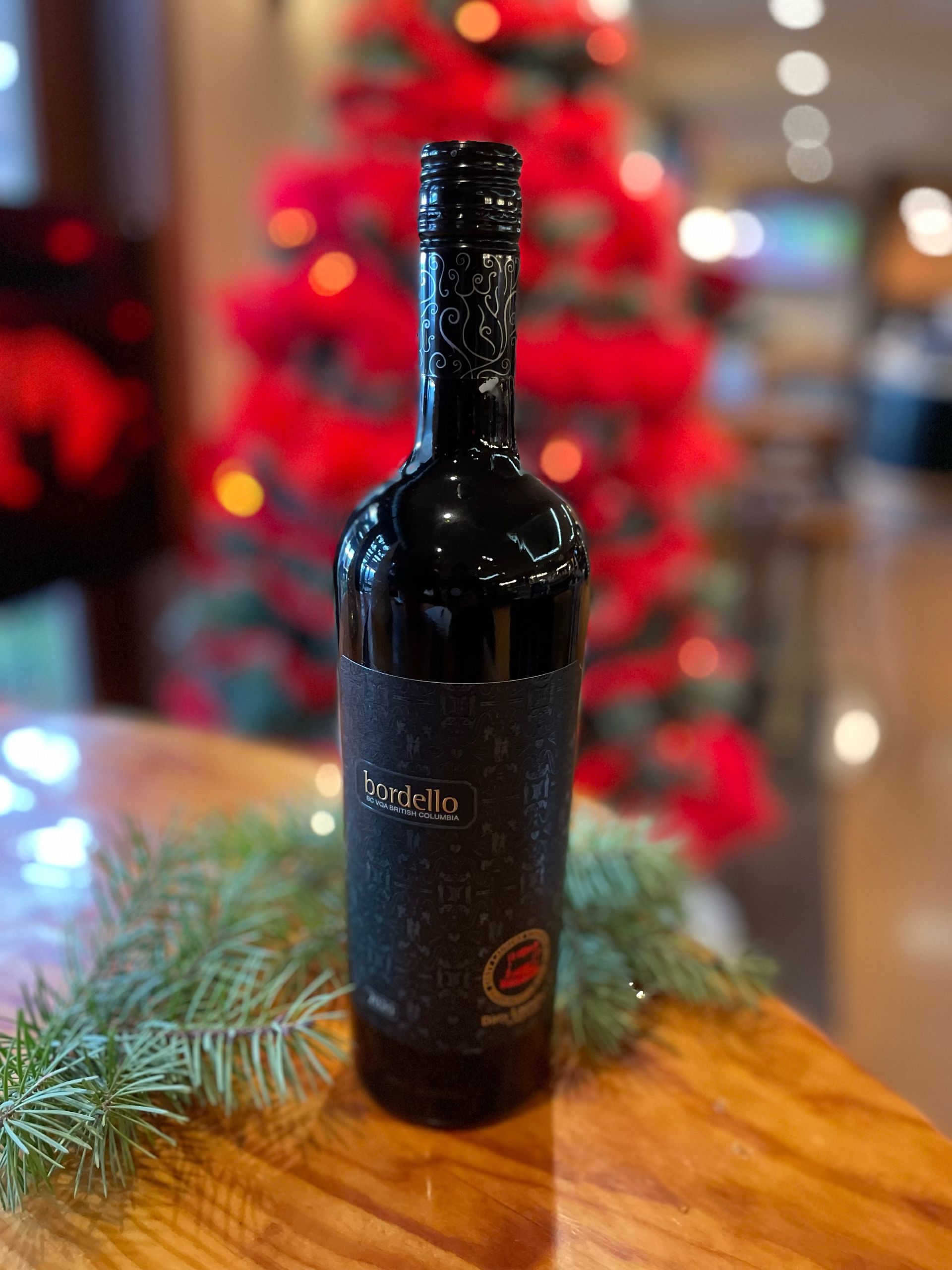 A bottle of wine is sitting on a wooden table in front of a christmas tree.