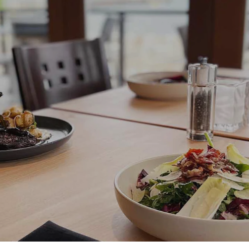 A bowl of salad sits on a table next to a plate of food