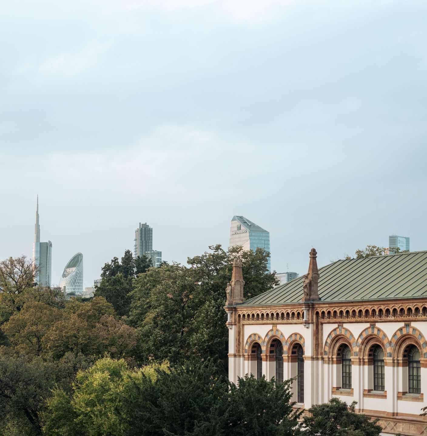Un grande edificio con il tetto verde è circondato da alberi e sullo sfondo si vede lo skyline della città.