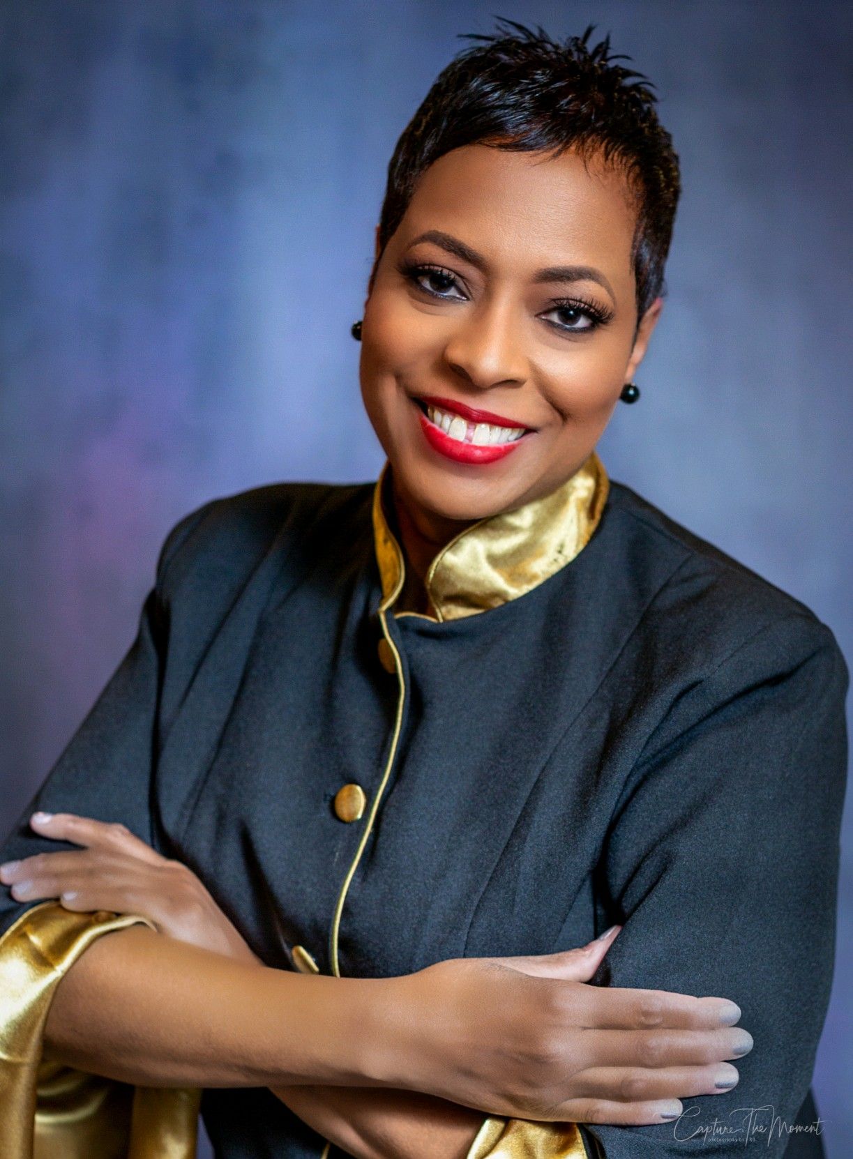 A woman in a black and gold jacket is smiling with her arms crossed.