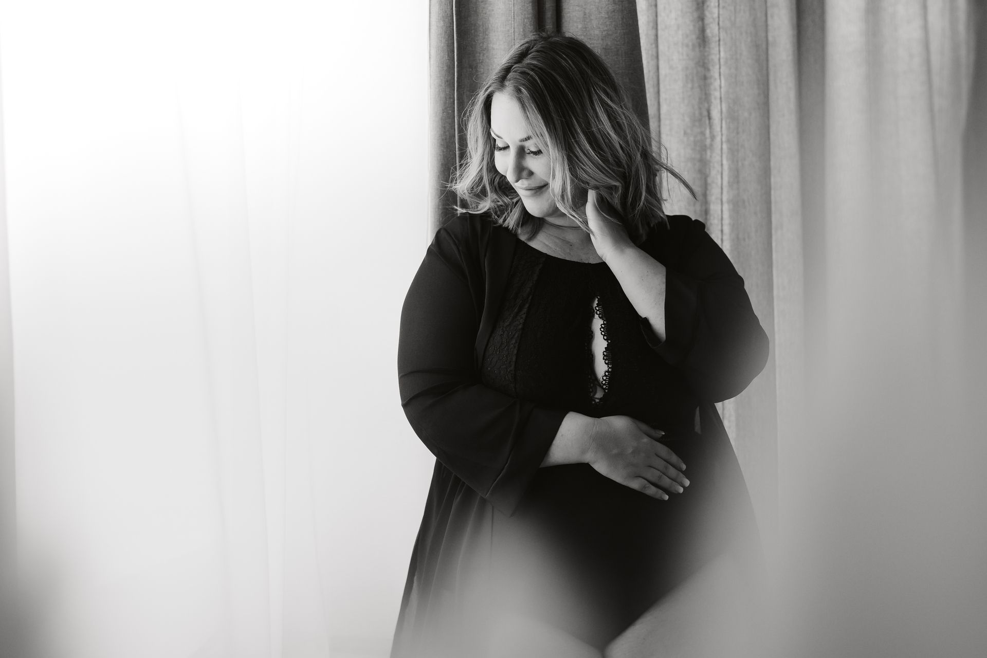 Eine schwangere Frau in einem schwarzen Kleid steht vor einem Fenster und hält ihren Bauch.